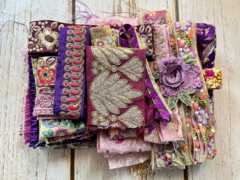 May include: Assortment of purple, pink, and gold fabric trims and embellishments. The trims feature floral and paisley patterns, and some have sequins or beads.