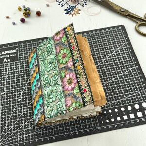 K&ouml;nnte beinhalten: Ein handgefertigtes Journal mit einem Stoffeinband, der vertikale Streifen aus bestickter Blumen-Spitze in Gr&uuml;n-, Rosa- und Goldt&ouml;nen aufweist. Das Journal hat dickes, strukturiertes Papier und liegt auf einer schwarzen Schneidematte mit Raster.