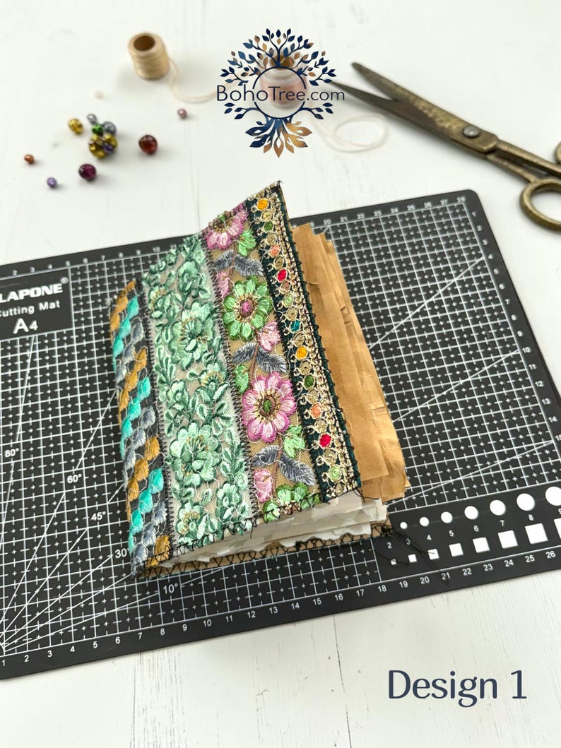 K&ouml;nnte beinhalten: Ein handgefertigtes Journal mit einem Stoffeinband, der vertikale Streifen aus bestickter Blumen-Spitze in Gr&uuml;n-, Rosa- und Goldt&ouml;nen aufweist. Das Journal hat dickes, strukturiertes Papier und liegt auf einer schwarzen Schneidematte mit Raster.