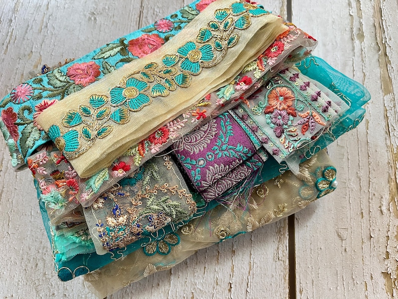 May include: A stack of colorful embroidered fabric trims with floral patterns in shades of teal, pink, gold, and purple. The trims are made of sheer and delicate materials, perfect for adding a touch of elegance to any project.