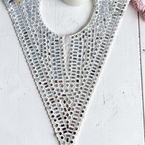 May include: A white, V-neck collar embellished with small, reflective mirror pieces. The collar has a scalloped trim and features the text "BohoTree.com". A lit candle and pink flowers are in the background.