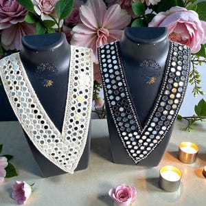 May include: Two decorative neckpieces on black mannequins. One is white with mirrored accents, the other is black with mirrored accents. The background features pink roses and candles, creating an elegant display.
