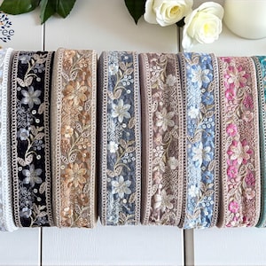 May include: An assortment of embroidered floral lace ribbons in shades of white, black, beige, blue, pink, and turquoise. The ribbons display detailed floral designs, ideal for crafts and decorative purposes. The word "Rose" is visible.
