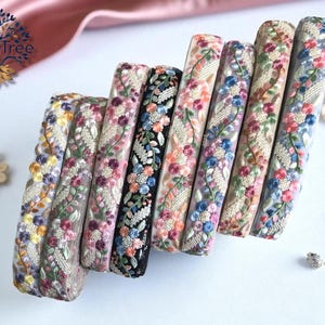 Pode incluir: Uma coleção de oito rolos de fita bordada com padrões florais intrincados em várias cores, incluindo rosa, azul, amarelo e verde, em diferentes tecidos de base como preto, bege e branco, dispostos em uma fileira diagonal.
