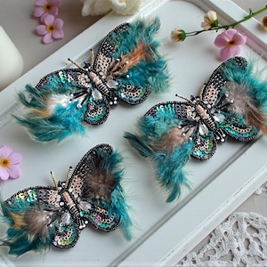 May include: Three decorative butterfly brooches with teal and brown feathers, sequins, and beadwork. The butterflies are arranged on a white tray with pink flowers. The brooches have a detailed design with a mix of textures and colors.