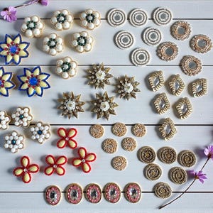 Può includere: Una collezione di vari abbellimenti fatti a mano, tra cui forme di fiori e stelle, disegni rotondi e ovali e dettagli a specchio. La tavolozza dei colori comprende bianco, oro, rosso e blu, disposti su una superficie di legno bianco.