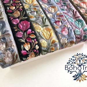 以下が含まれることがあります： グレー、黒、青など、さまざまな色の刺繍された花のリボンのクローズアップ。リボンは、ピンク、黄色、オレンジのアクセントが付いた詳細な花のパターンが特徴です。 Boho Treeのロゴが右下に表示されています。