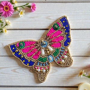 May include: A beaded butterfly decoration with pink, blue, green, and gold beads. The butterfly is embellished with small gemstones and pearls. The wings are detailed with intricate patterns. The decoration is placed on a white wooden surface, with flowers in the background.