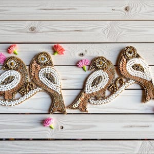 May include: Three decorative bird appliques with gold sequins, white beads, and brown accents are arranged on a white wooden surface. Small pink and yellow fabric flowers are scattered around the birds. The birds are perched on a branch.