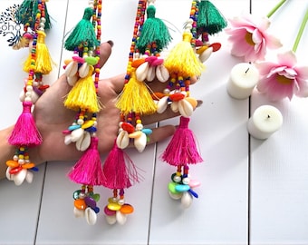 May include: Colorful beaded tassel garlands with yellow, green, and fuchsia tassels, adorned with seashells and vibrant beads. The garlands are displayed against a white background, with pink flowers and candles.