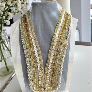 May include: A gold beaded and sequined collar necklace displayed on a mannequin. The necklace features a V-neck design with layers of gold beads, sequins, and small mirrored accents. The base is white mesh.