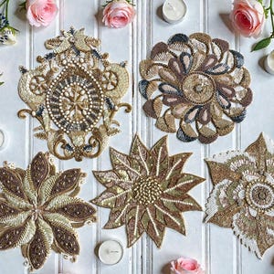 Könnte beinhalten: Draufsicht auf fünf verzierte, mit Perlen besetzte Tischsets in Gold-, Braun- und Cremetönen. Die Tischsets weisen aufwendige Blumen- und geometrische Designs auf. Rosa Rosen und Kerzen sind um die Tischsets herum angeordnet.