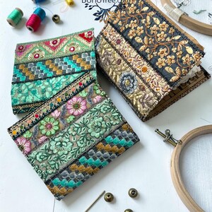 K&ouml;nnte beinhalten: Zwei quadratische Stoffnotizb&uuml;cher mit komplexen, mehrfarbigen floralen und geometrischen Stickmustern. Ein Notizbuch zeigt leuchtend gr&uuml;ne und rosa Blumenmotive, das andere goldene und schwarze Blumenstickereien. Enth&auml;lt den Text BohoTree.com.