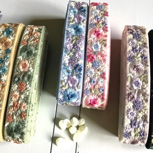 Puede incluir: Cinta floral bordada en varios colores. Las cintas presentan patrones florales detallados en tonos de azul, rosa, morado y verde sobre un fondo crema, azul claro y negro. Las cintas están apiladas y listas para usar.