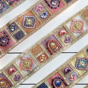 May include: Decorative fabric trim featuring a repeating pattern of embroidered squares in shades of pink, orange, purple, and gold. Each square contains intricate floral and geometric motifs outlined with metallic gold thread and sequins.