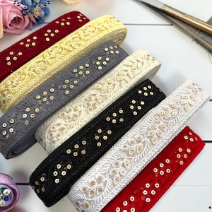 May include: Six rolls of decorative fabric trim arranged in a diagonal row. Each roll features a repeating embroidered leaf pattern accented with small, shiny gold sequins. The colors include deep red, bright yellow, dark gray, cream, black, and white.