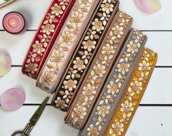 May include: Six rolls of decorative fabric trim arranged in a diagonal stack. Each roll features a repeating floral pattern embroidered with metallic gold thread on a base of red, white, black, beige, blue, and yellow fabric with sequins.