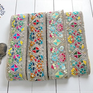May include: Close-up of four colorful embroidered ribbons with floral and geometric patterns. The ribbons feature a variety of colors including pink, blue, green, and yellow, with silver trim and sequins. A decorative metal clasp is visible on the left.