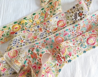 Puede incluir: Tres piezas de ribete bordado de colores vivos con diseños florales sobre fondo blanco. El ribete está hecho de una tela transparente con una textura delicada.