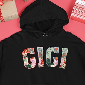 Op de afbeelding: Zwarte hoodie met de naam "GIGI" in een kleurrijk patchwork-ontwerp op de borst. De letters zijn gevuld met feestelijke patronen. De hoodie heeft een voorzak en een capuchon met trekkoord. De achtergrond is rood.