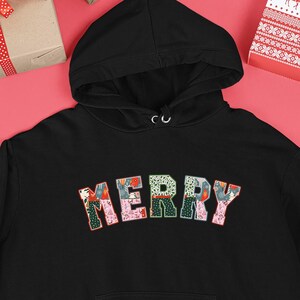 Op de afbeelding: Zwarte hoodie met het woord "MERRY" in kleurrijke patchwork-letters over de borst. De hoodie heeft een voorzak en een capuchon met trekkoord. De achtergrond bevat kerstversieringen en ingepakte cadeaus.