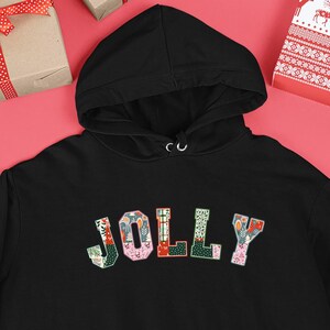 Op de afbeelding: Zwarte hoodie met het woord "JOLLY" in kleurrijke, patroonletters over de borst. De hoodie heeft een voorzak en een capuchon met trekkoord. De achtergrond bevat ingepakte cadeaus en kerstversieringen.