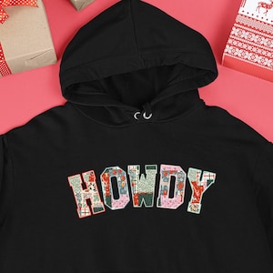 Op de afbeelding: Zwarte hoodie met het woord "HOWDY" in een kleurrijk, bloemmotief op de borst. De hoodie heeft een voorzak en een capuchon met trekkoord. De achtergrond toont ingepakte cadeaus en feestelijke decoraties.