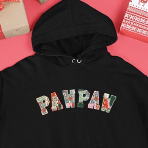 Op de afbeelding: Een zwarte hoodie met het woord "PAWPAW" in een kleurrijk patchwork-lettertype over de borst. De sweatshirt heeft een voorzak en een capuchon met trekkoord. De achtergrond bevat kerstversieringen en ingepakte cadeaus.
