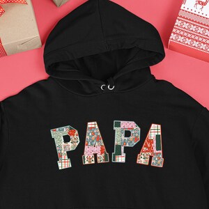 Op de afbeelding: Zwarte hoodie met het woord "PAPA" in een patchwork ontwerp. De letters zijn samengesteld uit verschillende patronen en kleuren. De hoodie heeft een voorzak en een capuchon met trekkoord. De achtergrond bevat ingepakte cadeaus en kerstversieringen.