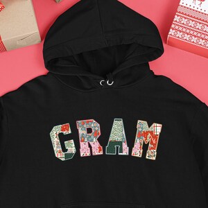 Op de afbeelding: Zwarte hoodie met het woord "GRAM" in een patchwork-ontwerp op de borst. De letters zijn gemaakt van verschillende stoffen met patronen in rood, groen en roze. De hoodie wordt omringd door ingepakte cadeaus en kerstversieringen.