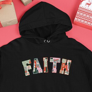Op de afbeelding: Zwarte hoodie met het woord "FAITH" in een kleurrijk patchwork-ontwerp op de borst. De hoodie heeft een voorzak en een capuchon met trekkoord. De achtergrond bevat ingepakte cadeaus en kerstversieringen.