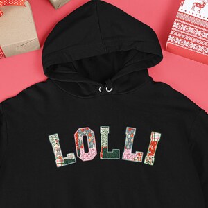 Op de afbeelding: Zwarte hoodie met het woord "LOLLI" in kleurrijke, patroonletters over de borst. De hoodie heeft een voorzak en een capuchon met trekkoord. De achtergrond bevat ingepakte cadeaus en kerstversieringen.