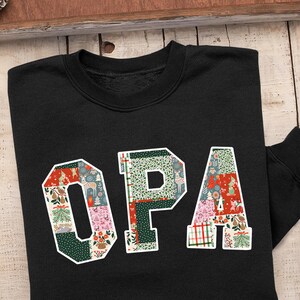 Peut inclure: Sweat-shirt noir avec le mot "OPA" en blanc, chaque lettre présentant un motif patchwork avec des motifs de vacances festifs. Le sweat-shirt est présenté sur une surface en bois avec des décorations de saison.
