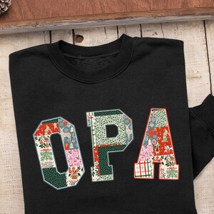 Peut inclure: Sweat-shirt noir avec le mot "OPA" dans un motif patchwork. Les lettres sont remplies de motifs festifs, notamment des arbres de Noël, des rennes et des motifs floraux. Le sweat-shirt est présenté sur une surface en bois.