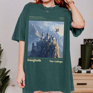 May include: Dark green t-shirt featuring a fantasy castle illustration with a flying dragon and the text "Basgiath War College". The shirt also includes the text "Threshing Day, October 1st, Only Dragon Riders Will Survive" and "Tiarendor Mavarre".