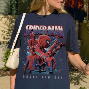 May include: Navy blue t-shirt with a Spider-Man graphic. The design includes multiple Spider-Man figures in red and blue, with the text "SPIDER-MAN" and "BRAND NEW DAY". The graphic is a collage of Spider-Man poses.