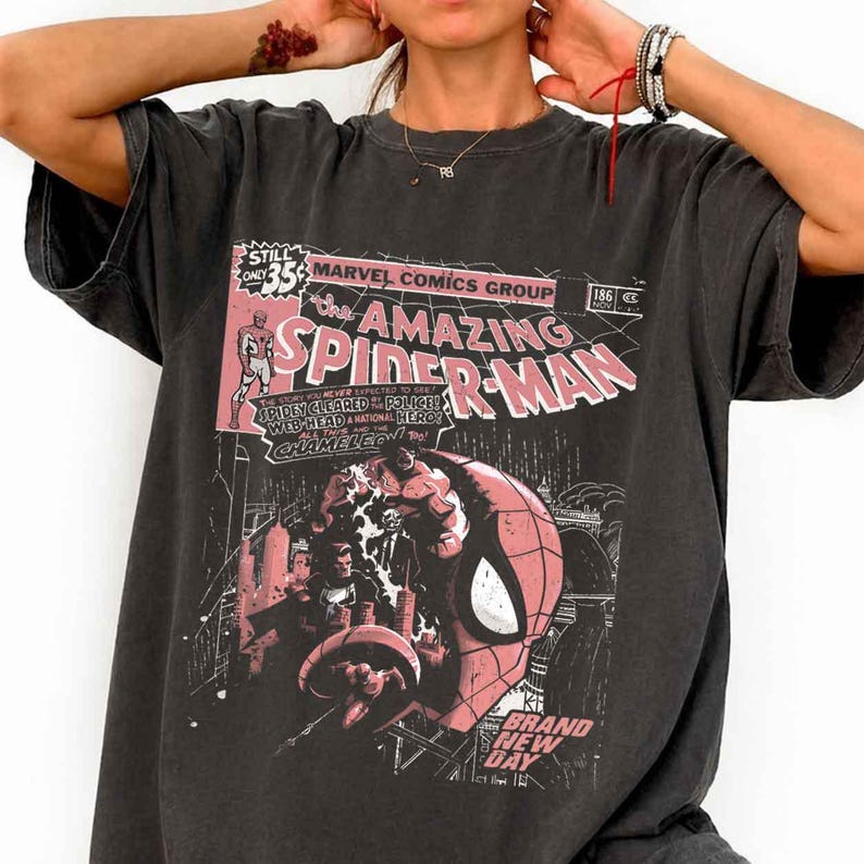 Puede incluir: Camiseta gris oscuro con un dise&ntilde;o de portada de c&oacute;mic vintage. El gr&aacute;fico incluye las palabras "Amazing Spider-Man" en rosa y blanco, detalles de c&oacute;mic y una ilustraci&oacute;n de Spider-Man. La camiseta tiene mangas cortas.