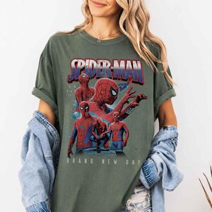 May include: An olive green t-shirt with a Spider-Man graphic. The design shows Spider-Man in different poses, with the text "SPIDER-MAN" in red and blue. Below the image is the phrase "BRAND NEW DAY". The shirt has short sleeves.
