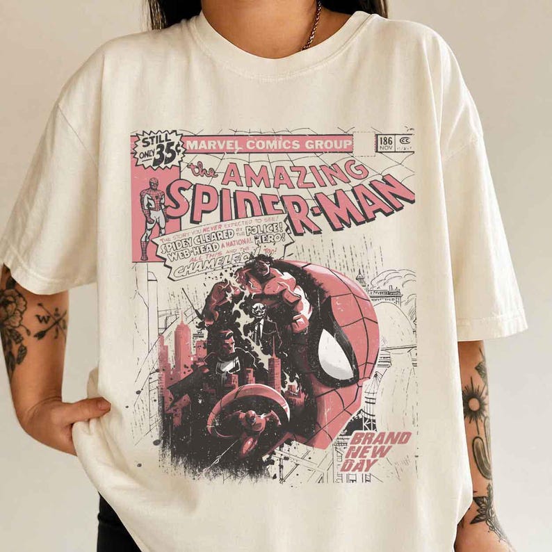 Puede incluir: Camiseta color crema con un dise&ntilde;o de portada de c&oacute;mic vintage de The Amazing Spider-Man. El gr&aacute;fico incluye el personaje de Spider-Man, paisajes urbanos y elementos de texto como "Marvel Comics Group" y "Brand New Day".