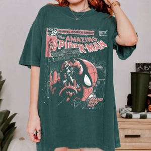 Puede incluir: Camiseta verde oscuro con un dise&ntilde;o de portada de c&oacute;mic vintage. El gr&aacute;fico incluye el texto "The Amazing Spider-Man" y una ilustraci&oacute;n de la cara de Spider-Man. La camiseta tiene un aspecto cl&aacute;sico y ligeramente descolorido.