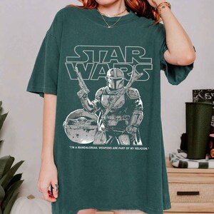May include: Dark green t-shirt featuring a Star Wars graphic. The design includes the Star Wars logo, a Mandalorian character with a weapon, and a depiction of Grogu. The text reads: "I'M A MANDALORIAN. WEAPONS ARE PART OF MY RELIGION."