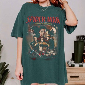 Op de afbeelding: Donkergroen T-shirt met een Spider-Man filmgrafiek. Het ontwerp bevat de woorden "Spider-Man" in het rood, met "Brand New Day" eronder. Het shirt toont een collage van personages uit de film.