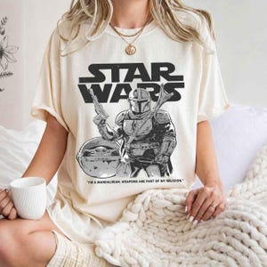 May include: Cream-colored Star Wars t-shirt featuring a black and white graphic of The Mandalorian holding a blaster, with Grogu in the background. The Star Wars logo is above the graphic, and the text "I'M A MANDALORIAN. WEAPONS ARE PART OF MY RELIGION." is below.