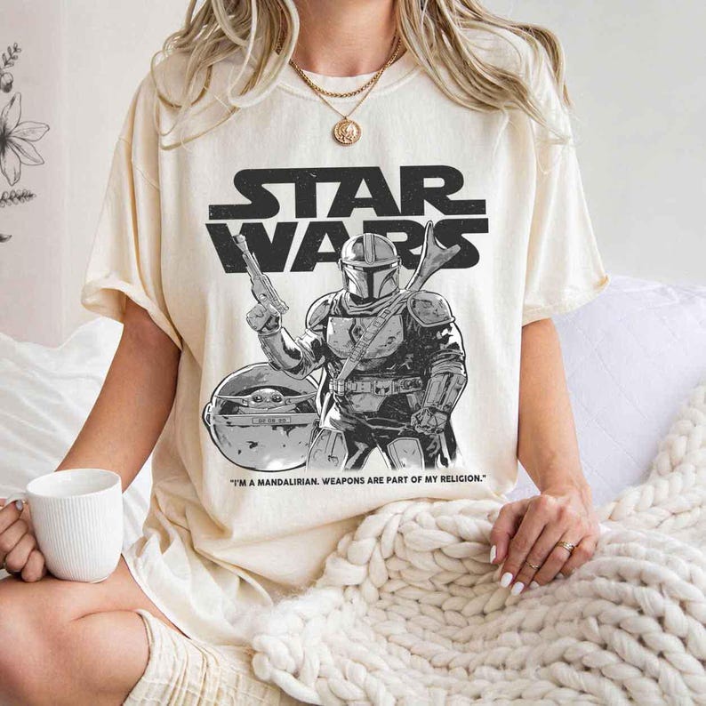 May include: Cream-colored Star Wars t-shirt featuring a black and white graphic of The Mandalorian holding a blaster, with Grogu in the background. The Star Wars logo is above the graphic, and the text "I'M A MANDALORIAN. WEAPONS ARE PART OF MY RELIGION." is below.