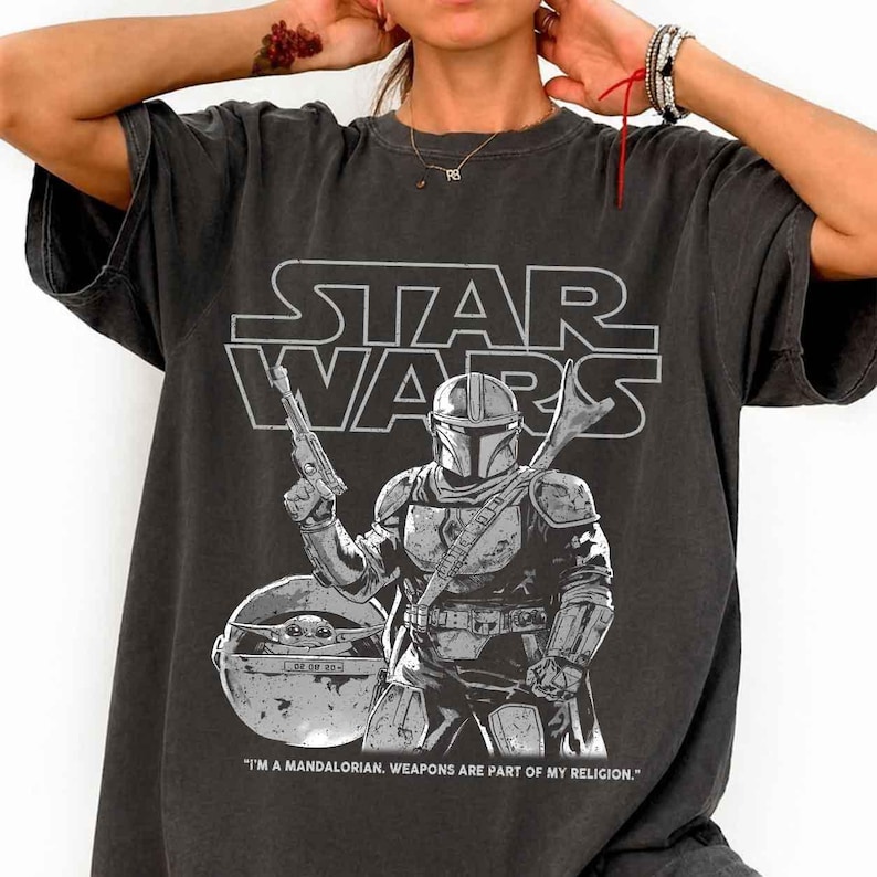 May include: Dark gray t-shirt featuring a Star Wars graphic. The design includes the Star Wars logo, a Mandalorian warrior with a blaster, and Grogu in his pod. The text reads, "I'M A MANDALORIAN. WEAPONS ARE PART OF MY RELIGION."