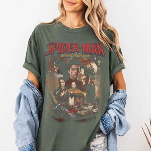 Op de afbeelding: Olijfgroen t-shirt met de rode tekst "SPIDER-MAN" en een afbeelding van personages uit de film. De tekst "BRAND-NEW DAY" staat onder de hoofdtitel. De mouwen zijn opgerold.