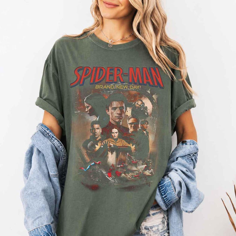Op de afbeelding: Olijfgroen t-shirt met de rode tekst "SPIDER-MAN" en een afbeelding van personages uit de film. De tekst "BRAND-NEW DAY" staat onder de hoofdtitel. De mouwen zijn opgerold.