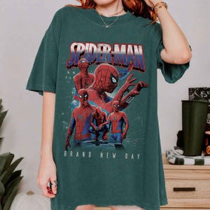 May include: Dark green t-shirt with a Spider-Man graphic. The design features Spider-Man in different poses, with the text "SPIDER-MAN" in red and blue, and "BRAND NEW DAY" in white. The shirt has short sleeves and a loose fit.