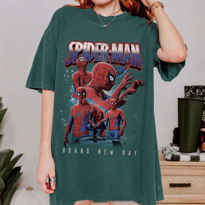 May include: Dark green t-shirt with a Spider-Man graphic. The design features Spider-Man in different poses, with the text "SPIDER-MAN" in red and blue, and "BRAND NEW DAY" in white. The shirt has short sleeves and a loose fit.