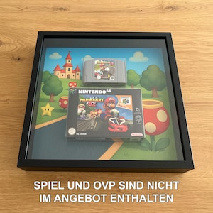 Könnte beinhalten: Gerahmte Nintendo 64-Videospiel-Präsentation mit einer Mario Kart 64-Spielbox und einer Mario Party X-Cartridge. Der Hintergrund zeigt eine farbenfrohe Szene mit Bäumen, einem Schloss und Pilz-Grafiken. Der Rahmen ist schwarz.